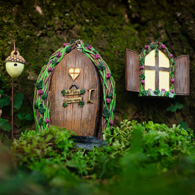 Hand-painted opening fairy door and window set mounted on a garden tree at dusk with glowing windows
